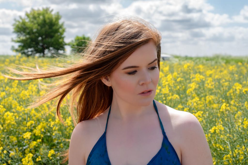 A fully edited portrait image of a redhead woman in a summer dress standing in a field of rapeseed flowers